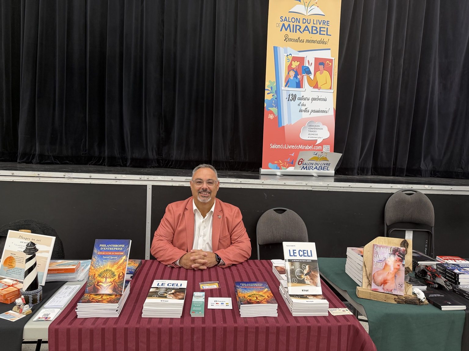 Patrick Charette assis derrière son kiosque au Salon du livre de Mirabel, avec ses livres et la bannière officielle en arrière-plan.